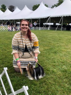 Woman kneeling on grass with a small dog on a leash, in front of white tents at an outdoor event.