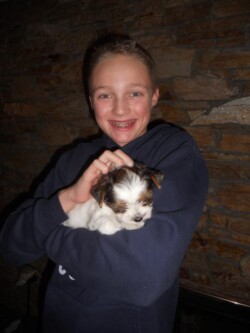 A young person with braces smiles while holding a small white and brown puppy against a stone wall background.